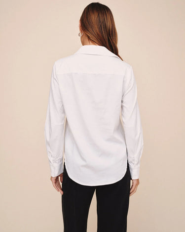 A woman viewed from the back wearing a white long-sleeve button-up shirt and black pants, standing against a plain beige studio background.