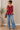 Woman wearing a red sleeveless top and blue jeans standing indoors.
