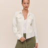 A woman modeling a light cream-colored, long-sleeve button-up shirt jacket over a white top and olive green mini skirt, set against a solid beige background.