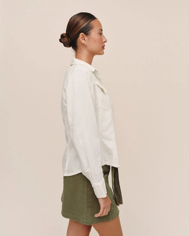 A woman in side profile wearing a white long-sleeve button-up shirt and an olive green mini skirt, standing against a solid light beige background.