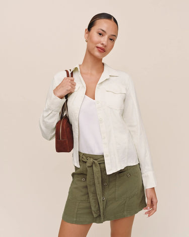 A woman modeling a white long-sleeve button-up shirt over a white top, paired with an olive green belted mini skirt and carrying a brown shoulder bag, set against a solid beige background.