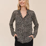 A woman modeling a black long-sleeve button-up blouse with a small white floral or ditsy print, paired with black pants, against a solid beige background.