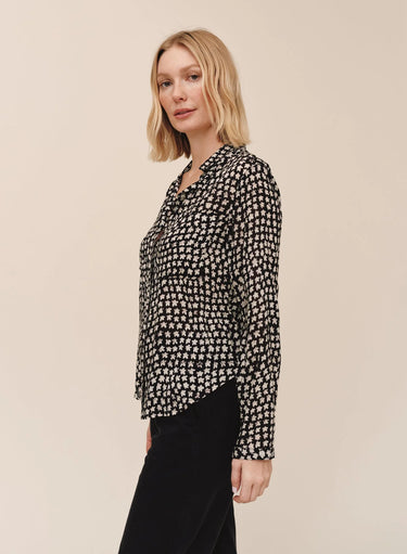 A woman with blonde shoulder-length hair is shown in profile wearing a black long-sleeve button-up blouse with a dense white floral or ditsy print, paired with black pants, against a solid light beige studio background.