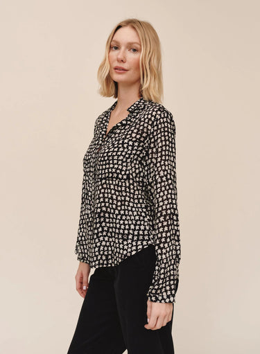 A blonde woman wearing a black long-sleeve button-up blouse with a small white geometric pattern, paired with black pants, standing against a solid beige studio background.