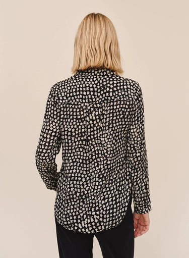 A person viewed from the back wearing a black long-sleeve button-up shirt with a dense small floral or ditsy print in a warm off-white/cream tone, paired with black pants, against a plain beige studio background.