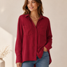 A woman wearing a long-sleeve button-up shirt in deep burgundy with two chest pockets, paired with blue jeans, standing against a warm beige background.