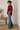 A woman stands in profile wearing a burgundy button-up shirt and blue flare jeans, standing on a woven jute rug in a bright room with a wooden bench and vase on the left.
