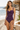 A woman modeling a ruched one-piece swimsuit in deep purple on a sandy beach with the ocean and a thatched umbrella in the background.