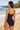 A woman wearing a black one-piece swimsuit stands on a sandy beach with the ocean and rocks in the background, photographed from behind.