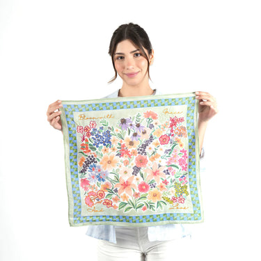 Woman holding a colorful green floral scarf with a white background with embroidered words “ Bloom with Grace and Love” in four corners of scarf.