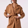 A woman wearing a camel brown belted trench coat with shoulder epaulets, paired with brown trousers, against a plain light background