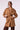 A woman wearing a camel brown belted trench coat with shoulder epaulets, paired with brown trousers, against a plain light background