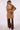 A woman wearing a camel brown belted trench coat with shoulder epaulets, paired with brown trousers, against a plain light background