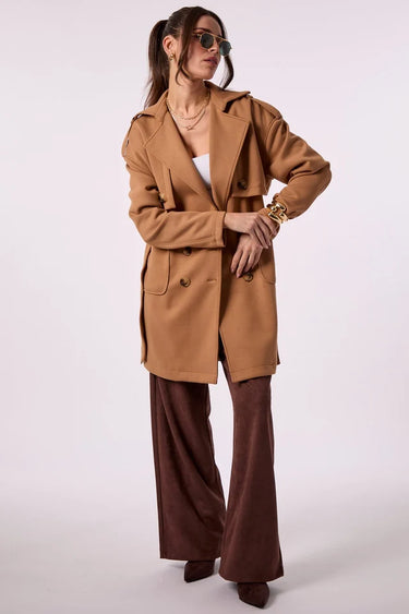 A woman wearing a camel brown belted trench coat with shoulder epaulets, paired with brown trousers, against a plain light background