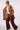 A woman wearing wearing a camel brown belted trench coat with shoulder epaulets, opening the coat to show the plaid lined interior paired with brown trousers, against a plain light background