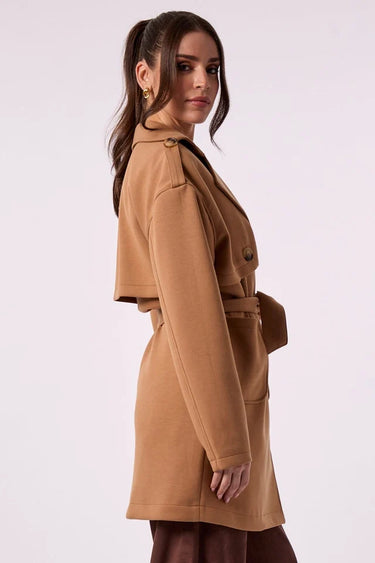 A model standing sideways wearing a camel brown belted trench coat with shoulder epaulets, paired with brown trousers, against a plain light background