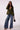 A woman models an olive green blazer over a matching green top, paired with wide-leg blue jeans, brown heels, and a small brown handbag against a plain light background.