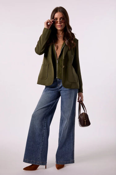 A woman models an olive green blazer over a matching green top, paired with wide-leg blue jeans, brown heels, and a small brown handbag against a plain light background.