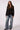 A woman modeling a black blazer over a black top with wide-leg blue jeans, standing against a light gray studio background.