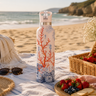 Beach picnic setup with a water bottle, sunglasses, and food on a blanket.
