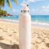 Pink Palm patterned water bottle with a silver cap on a sandy beach with ocean and sky in the background.