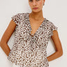 woman wearing flowy blouse in espresso daisy print with white pants on a white background