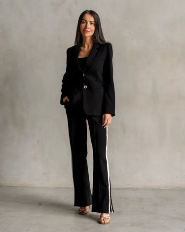 a woman wearing a black blazer with gold button accents and black pants with a white stripe on a neutral background