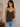 Woman wearing a dark gray tank top and blue jeans indoors.