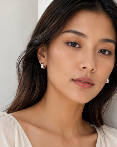 A close-up portrait of a woman wearing small silver hoop earrings with clear gemstones, set against a soft neutral background.