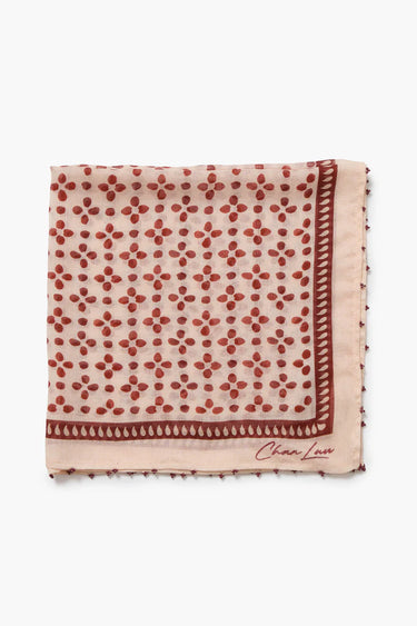 A square scarf in a light peach-beige color with a repeating rust-red floral pattern and a matching rust-red border, featuring the 'Chanel' logo embroidered in the bottom right corner, set against a plain white background.