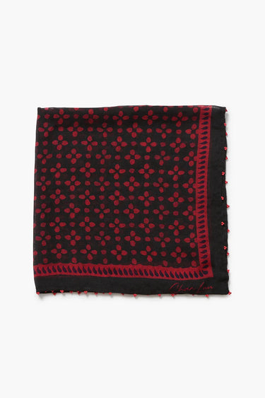 A square black scarf with a red geometric pattern, featuring a red border with small pom-pom details and a signature logo in the bottom right corner, displayed flat against a light gray background.