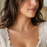 A woman wearing a delicate silver necklace with a small pendant, shown in a close-up portrait. She is wearing a cream-colored ruffled top.