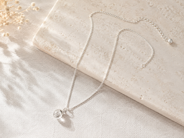 A delicate silver chain necklace with a clear glass bead pendant, styled on a textured cream stone slab with dried floral accents.