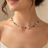 A close-up of a woman wearing a delicate multi-colored gemstone choker necklace with a gold chain, set against a soft beige background.