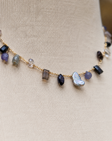 A gold chain necklace with irregular gemstone beads in purple, gray, black, and a large white mother-of-pearl heart, displayed on a beige fabric mannequin bust.