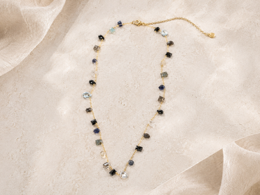 A delicate gold chain necklace with small, irregular gemstone beads in dark blue, black, gray, and clear, arranged on a draped beige fabric background.