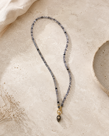 A beaded necklace with gray and black faceted beads, a gold-toned ring connector, and a dark brown teardrop pendant, displayed on a textured beige stone surface.