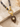 A close-up of a gold-tone pendant necklace featuring a carved dark brown leaf charm with a clear round stone, attached to a gold ring and a beaded strand with gray and navy beads, styled on a textured stone slab and beige fabric.
