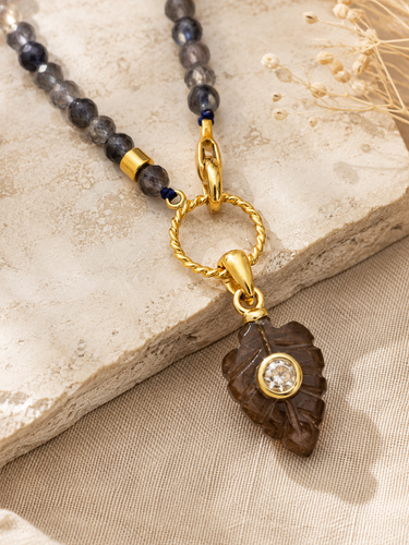 A close-up of a gold-tone pendant necklace featuring a carved dark brown leaf charm with a clear round stone, attached to a gold ring and a beaded strand with gray and navy beads, styled on a textured stone slab and beige fabric.