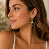A woman in profile wearing a gold horsebit drop earring, shown in a warm, softly lit indoor setting.