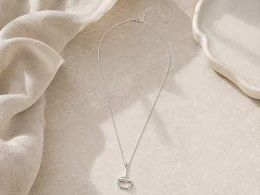 A delicate silver necklace with a small silver pendant, styled on a textured beige surface with draped cream fabric and a white ceramic dish.