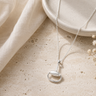 A silver pendant necklace with a minimalist ring and teardrop design, displayed on a textured beige stone surface next to a cream linen cloth and dried flowers.