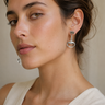 A close-up of a woman wearing a silver drop earring with a rectangular clear stone and an oval dangling element, paired with a cream-colored top against a neutral beige background.
