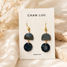 Chan Luu drop earrings with dark gray and navy blue stone discs on gold hooks, displayed on a white brand card with dried flowers and textured stone props.