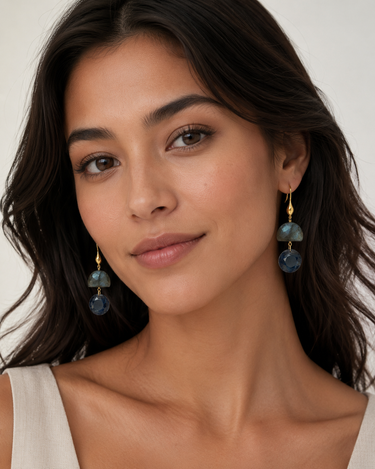 A close-up of a woman wearing gold dangle earrings with stacked round blue-green stone beads, shown against a light neutral background.