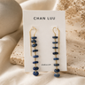 A pair of Chan Luu dangle earrings with dark blue stone beads and gold-toned metal, displayed on a white brand card against a warm beige fabric and stone background.