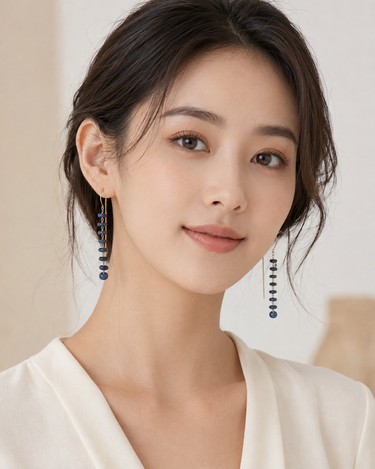 A close-up portrait of a woman wearing long, dangling earrings with dark blue bead-like drops and gold-toned chain details, paired with a cream-colored top against a soft beige background.