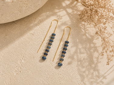 A pair of gold drop earrings with small dark blue beads, styled on a textured beige surface with soft shadows and dried floral decor.