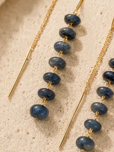 Gold-toned beaded necklace with dark blue stone beads and a long straight gold bar, shown on a light beige textured surface.