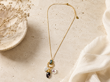 A delicate gold chain necklace with a multi-stone pendant, styled on a textured beige stone surface next to a small ceramic dish with dried flowers and a white fabric.